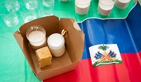 De la nourriture traditionnelle haïtienne dans une boîte en carton à emporter. À côté, sur la table, se trouve le drapeau haïtien.