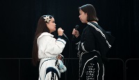 Two young women in traditional Inuit attire are holding microphones and facing each other