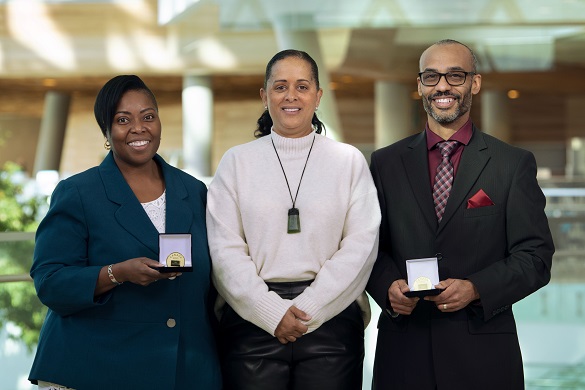 Three people standing next to each other and smiling. Two of them are holding awards