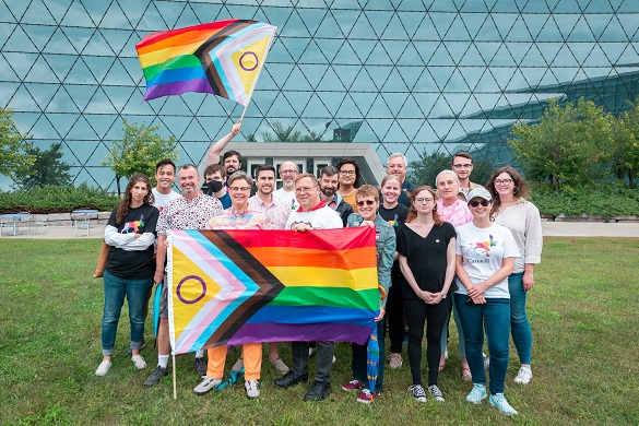 A group of people gathered together outside. Two of them are holding the inclusive Pride flag.