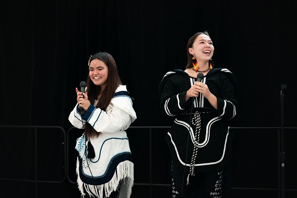 Two young women in traditional Inuit attire are holding microphones and smiling